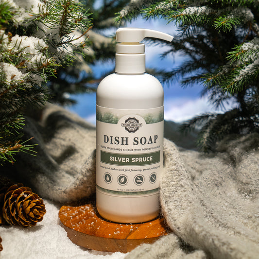 Rustic Strength, Dish Soap, 16oz, White Pump bottle, Silver Spruce photoshoot image set on a wooden plate with knit fabric behind surrounded by snow covered evergreen branches and pinecones