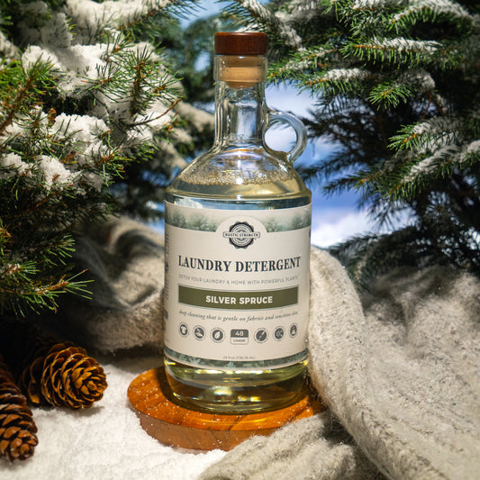 Rustic Strength, Laundry Detergent, 24oz, Glass Bourbon Bottle, Silver Spruce, photoshoot image with bottle set on a wooden plate in the snow with a knit fabric behind, surrounded by evergreen boughs and pine cones.