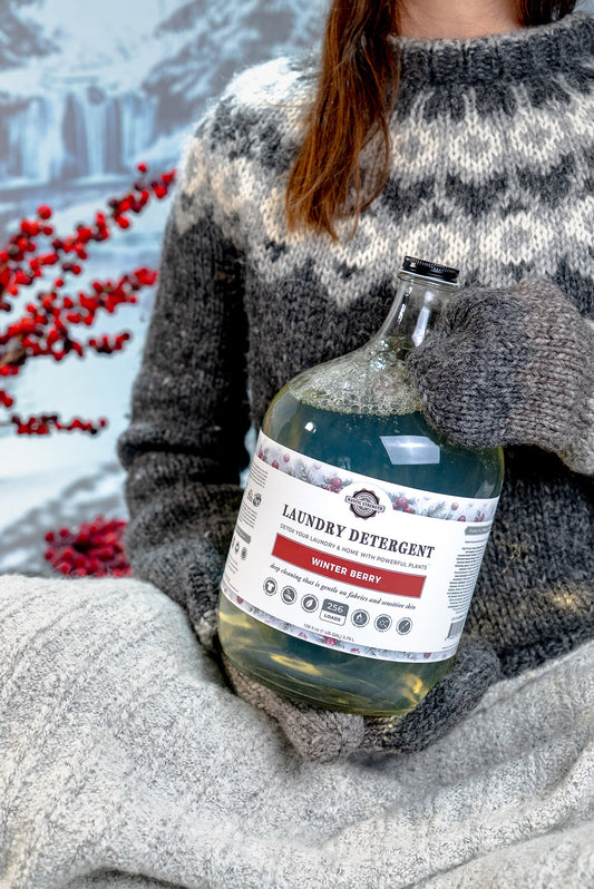 Rustic Strength, person wearing a gray knit sweater and matching knit gloves holds a bottle of laundry detergent against a softly blurred outdoor winter background with red berries.