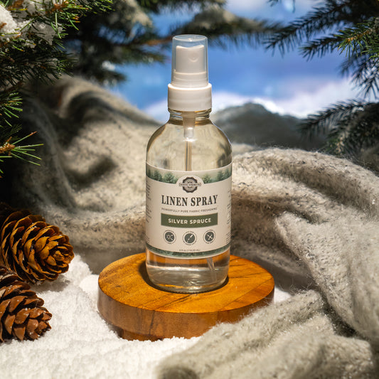 Rustic Strength, Linen Spray, 4oz, Glass with spray top, Silver Spruce, photoshoot image set on a wooden plate, set in the snow in front of some knit fabric, surrounded by snow covered evergreen branches and pinecones